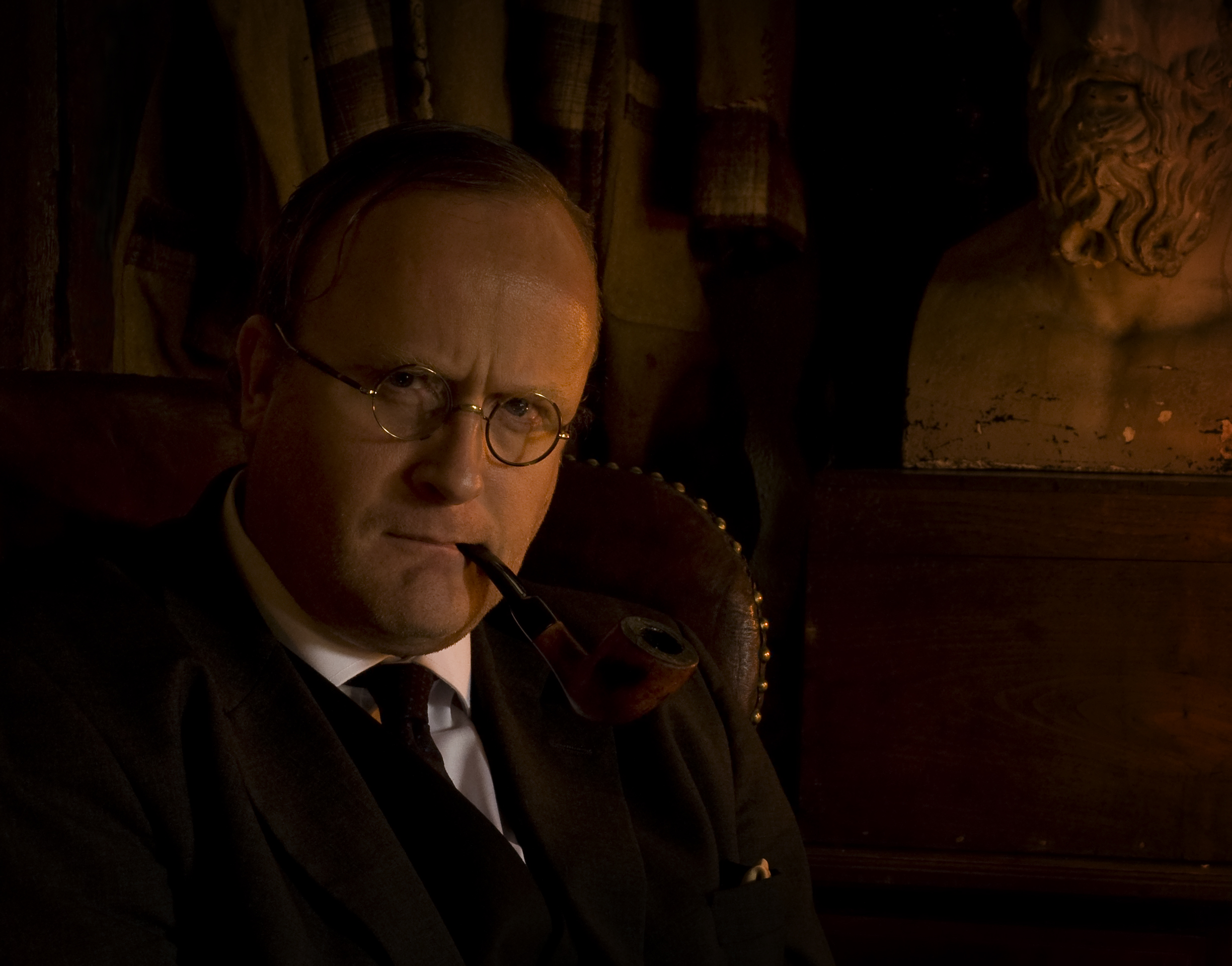 A person, smoking a pipe, sits in a leather chair in a darkened room.