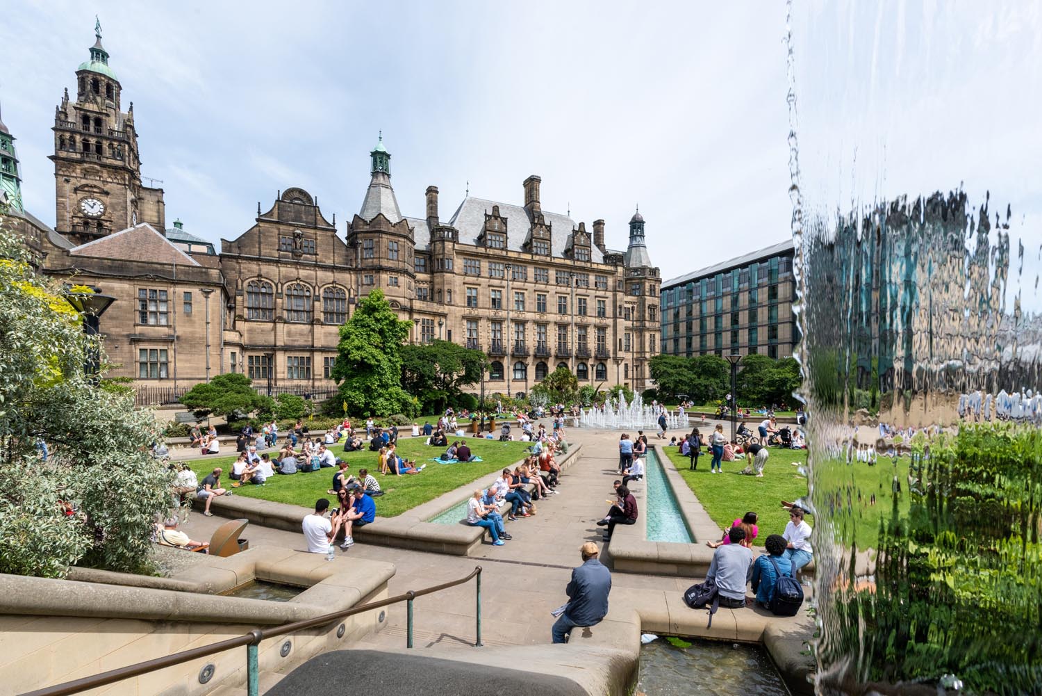 A view of the Peace Gardens on a sunny day. People are walking about while others are sitting on the grass. In the background is Sheffield Town Hall.