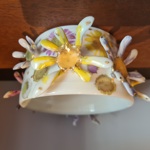 Close-up of a handmade ceramic bowl displayed on a wooden surface. The white bowl is decorated with colourful floral patterns and three-dimensional flower shapes attached around the outside. Yellow, purple and white petals with gold accents protrude from the surface, creating a textured, sculptural design. The background is softly blurred, drawing attention to the intricate craftsmanship.