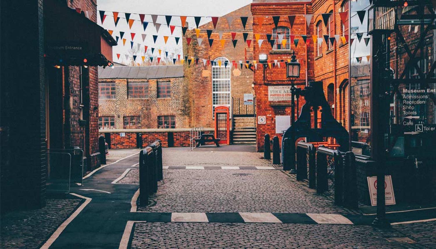 Cobblestone courtyard surrounded by historic brick buildings, decorated with strings of triangular bunting in red, white, and blue. A traditional red phone box stands near a large arched window at the far end. Signs on the right indicate museum entrance, admissions, shop, café, and toilets.