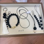 Close-up of a jewellery and accessories display in a wooden tray lined with cream fabric and branded “oicé.” The collection includes a black-and-cream tassel keyring, a beaded bracelet with a gold clasp, a long cord necklace with black and beige beads, and two beaded bag charms or phone straps in matching monochrome tones. The tray is photographed against a light wood floor, highlighting the minimalist handmade design.
