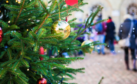 Close-up of a decorated Christmas tree with green branches adorned with shiny ornaments, including a gold bauble reflecting the surroundings and smaller red baubles. In the blurred background, people are walking on a cobblestone street near a building, suggesting a festive outdoor setting.