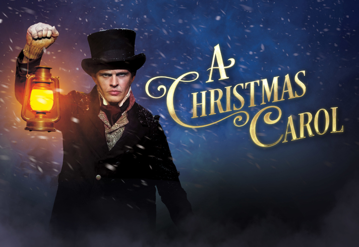 Promotional image for A Christmas Carol. A person in Victorian-style clothing and a tall black top hat stands in a snowy, dark blue scene, holding a glowing lantern that casts warm light across the image. Snow swirls through the air and mist fills the foreground. Large gold lettering on the right reads “A Christmas Carol,” creating a dramatic, atmospheric festive theatre poster.