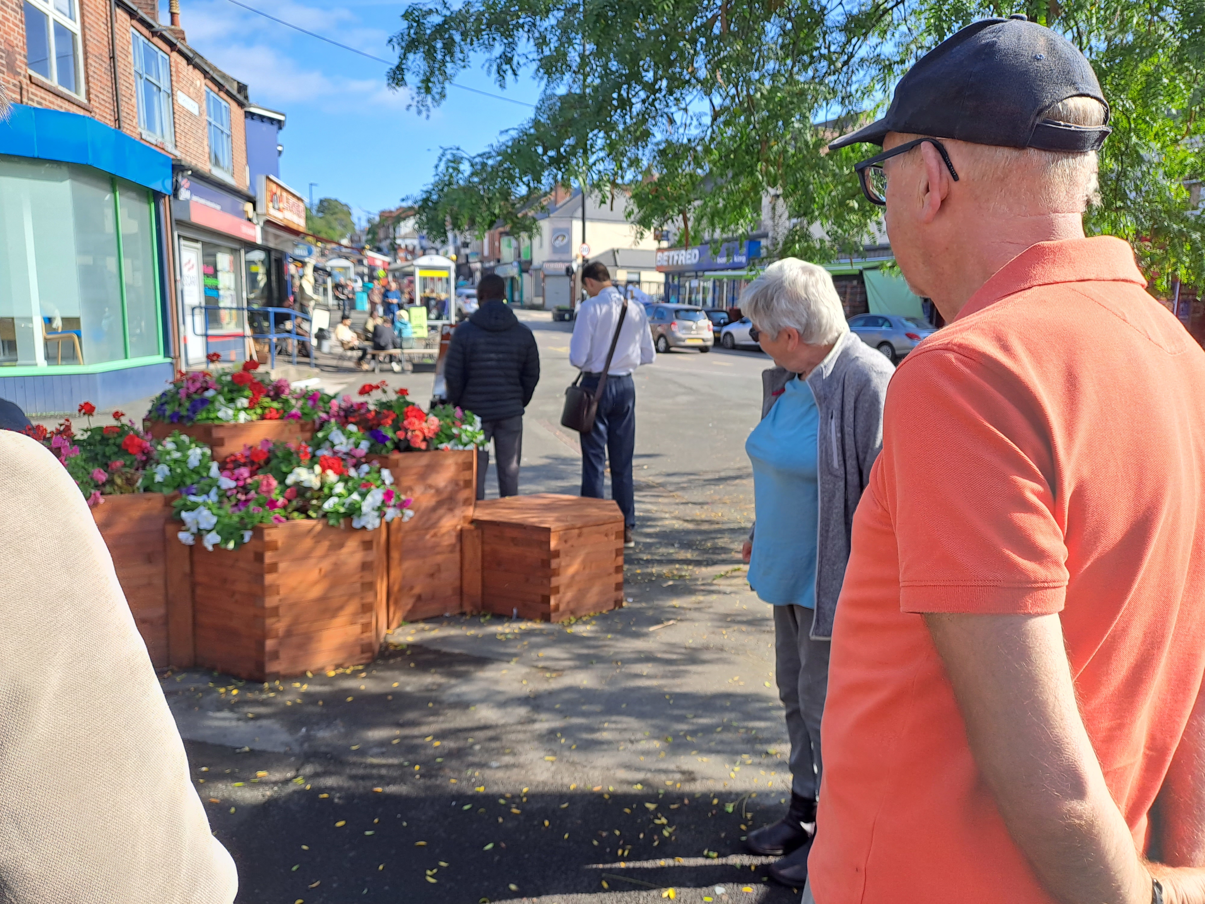 Street scene with several people standing near a group of wooden planters filled with colorful flowers. The planters are arranged in a stepped formation on the pavement, and shops line the street in the background under a clear blue sky.