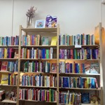 A set of tall wooden bookshelves filled with brightly coloured books arranged neatly across multiple rows. Warm lighting highlights sections of the shelves, and small decorative items, such as dried flowers and framed prints, sit on the top shelf. A woven lamp glows softly on the right-hand side.