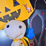 Photograph of Halloween-themed crafts displayed on a dark surface. A large yellow paper jack-o’-lantern face decorated with blue gems and animal stickers hangs in the background. In the foreground is a handmade white ghost figure with googly eyes, a string smile, yellow ribbon and colourful beads. Around it are coloured feathers, jewel stickers, craft sticks, and small animal character cut-outs, with chalk-drawn shapes visible on the black background.