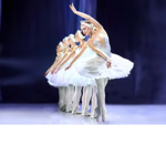 A line of ballet dancers in white tutus and matching headpieces perform on stage. The dancers stand in a diagonal formation, with the nearest dancer extending one arm overhead and leaning gracefully forward. Soft blue and purple stage lighting illuminates the scene, while dark curtains and a blurred stage backdrop create depth behind the performers.