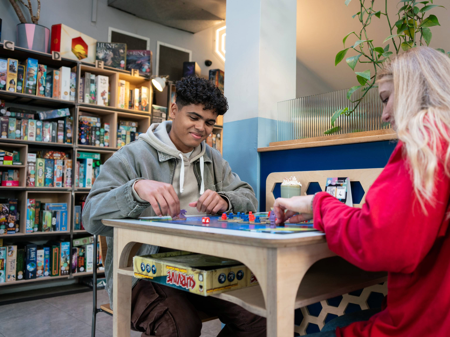 Two people are sat at a table, in a cafe, playing a board game and drinking coffees. Behind them is shelving stacked full of all different board games.