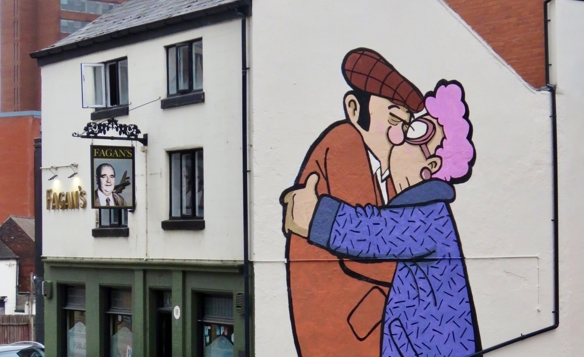 Street art mural on the side of a white building depicting two cartoon-style characters embracing, one wearing an orange coat and flat cap and the other in a purple coat with curly pink hair. The artwork is signed ‘McKee’ and located above a pub called Fagans.