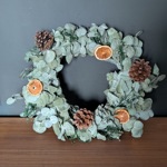 A circular wreath made of dried eucalyptus leaves, accented with pinecones and slices of dried orange. The wreath is displayed against a dark grey wall on a wooden surface, creating a rustic and natural look.