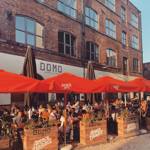 An outdoor dining area in front of a brick building with large windows and a sign reading “DOMO.” The seating area is covered by bright red umbrellas labeled “Aperol Spritz” and surrounded by wooden planters filled with greenery. Several people are seated at wooden tables enjoying food and drinks. The scene is lit by warm sunlight, creating a lively and inviting atmosphere.