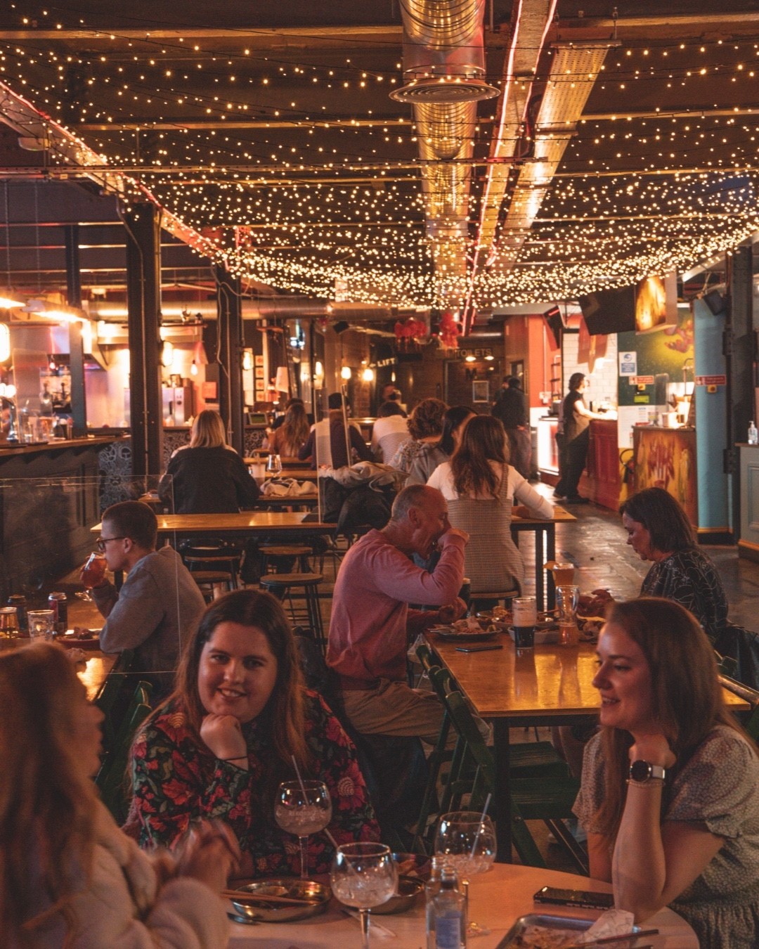 A view inside Cutlery Works: there are people sat at tables eating, chatting and having a good time. The ceiling is festooned with twinkling fairy lights. 