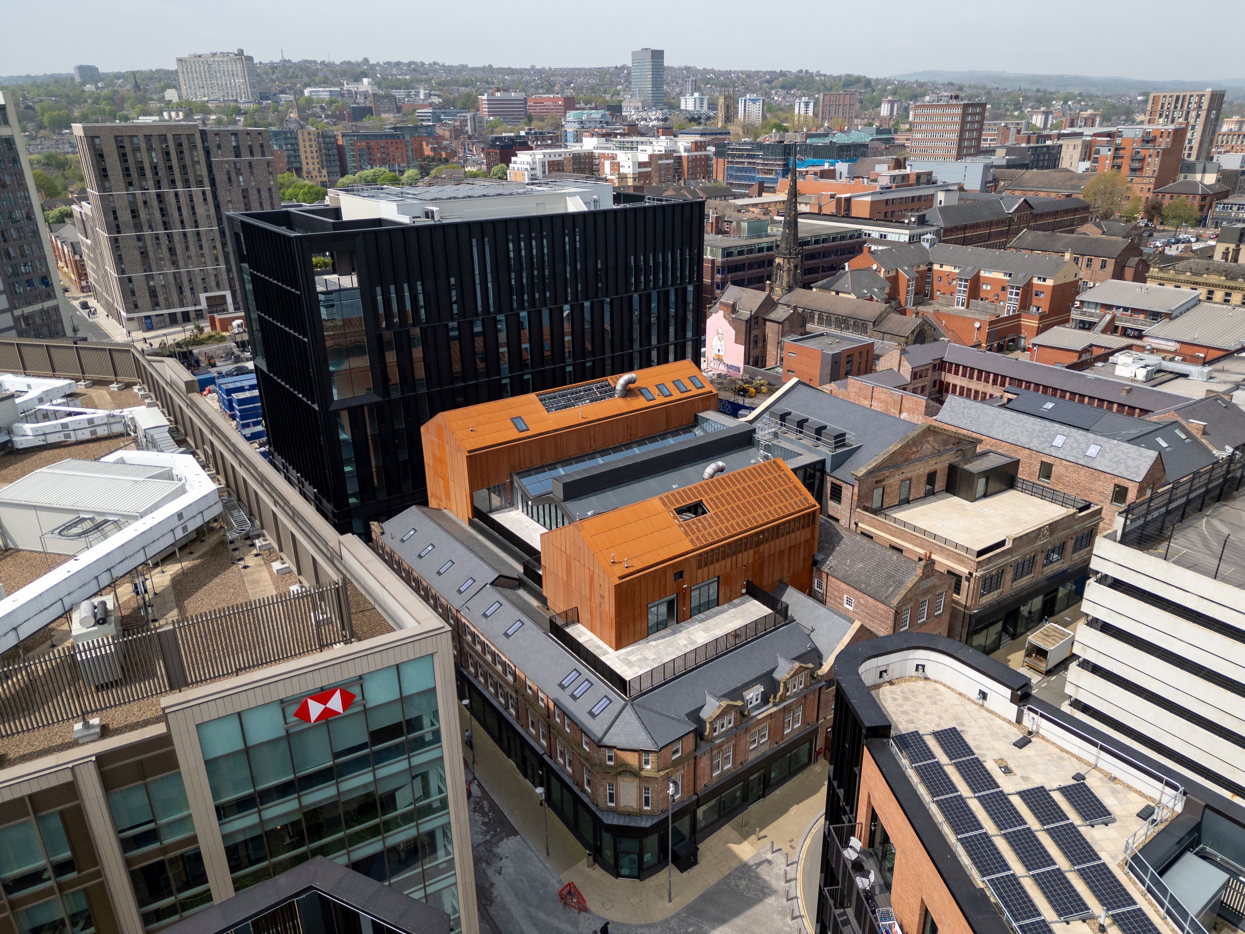 Aerial view of an urban area featuring modern and historic architecture. Prominent buildings include a black glass high-rise and orange-clad structures surrounded by older brick buildings. The scene shows a mix of commercial and residential properties, with solar panels visible on one rooftop and an HSBC building in the foreground. The background includes a sprawling cityscape with trees and hills under a clear sky.