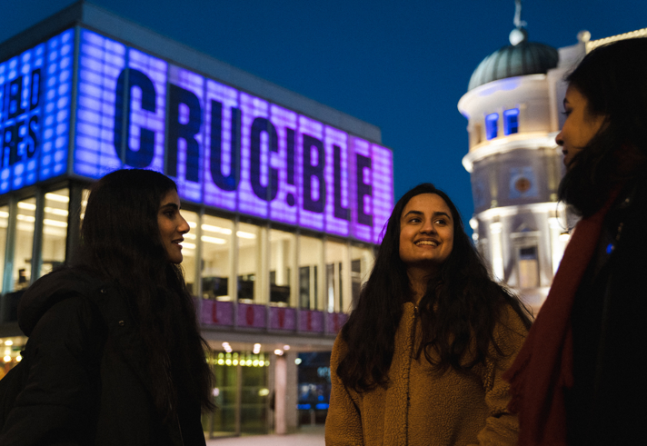 Khyati Mundrey Srivastav - University of Sheffield student: Three people standing outside the Crucible Theatre at night, with the building illuminated in purple and blue lights displaying the word “CRUCIBLE.” A historic domed building is visible in the background under a dark sky.