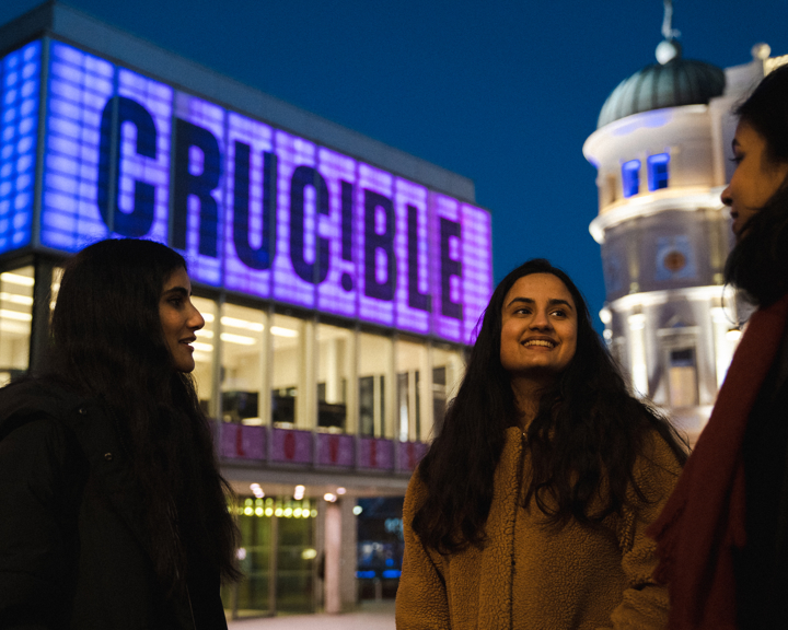 Khyati Mundrey Srivastav - University of Sheffield student: Three people standing outside the Crucible Theatre at night, with the building illuminated in purple and blue lights displaying the word “CRUCIBLE.” A historic domed building is visible in the background under a dark sky.