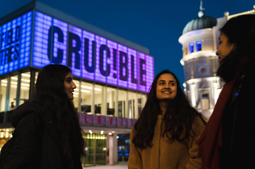 Three people standing outdoors in front of the Crucible Theatre at night. The building is brightly lit with a large illuminated sign reading “CRUCIBLE” in purple and white. Another building with a dome and ornate architecture is visible in the background under a dark blue sky.