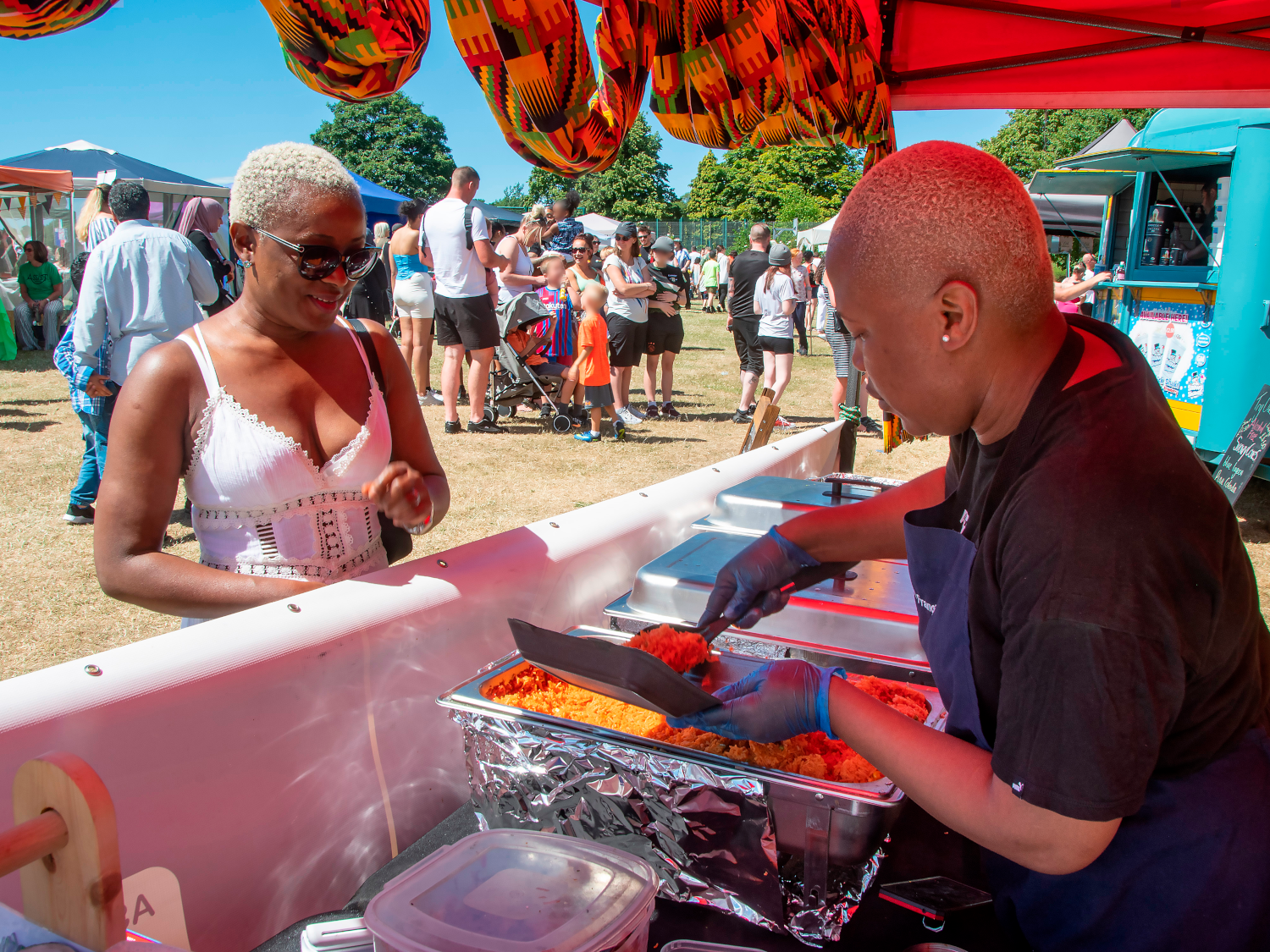Food stall at an outdoor event with a person serving rice from metal trays under a red canopy decorated with patterned fabric. A crowd of people, including families with children and strollers, is visible in the background on a grassy area with trees and a blue food truck.