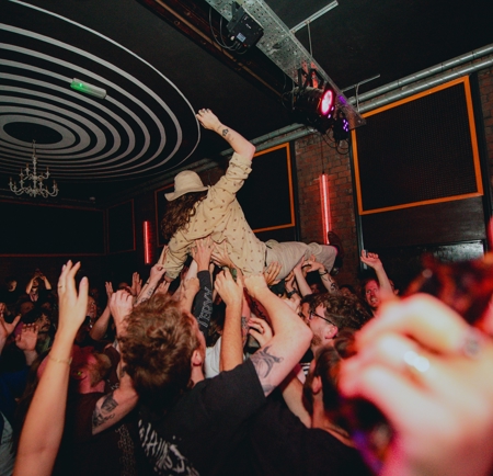 A crowd dances at an intimate gig, with one person crowd-surfing across the top of the audience.