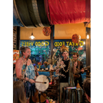 Live music performance inside a cosy café decorated with colourful hanging fabrics, flowers and warm lighting. Three performers stand with microphones and instruments, including a hand drum and acoustic guitar, in front of a window painted with the words “Great Food” and “Good Vibes.” Audience members are visible in the foreground, while decorative branches and lamps create an intimate, bohemian atmosphere.