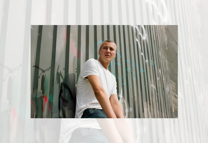 A person in a plain white T-shirt and jeans sits leaning against a corrugated metal wall covered with graffiti. The photograph is taken from a low angle, with the vertical lines of the grey metal panels stretching into the background. Bright daylight illuminates the scene, creating strong contrast between the white clothing and the industrial urban setting. The person looks off to one side while seated against the wall.