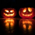 Two carved Halloween pumpkins glow orange in the dark, lit from within by candles or lights. The pumpkin on the left has sharp, slanted eyes and a mischievous grin, while the pumpkin on the right features a wide smile and large eyes. Both sit on a glossy surface that reflects their warm light. The background is almost entirely black, creating a spooky atmosphere that highlights the illuminated jack-o'-lantern faces.  Image by David Menidrey - Unsplash images website