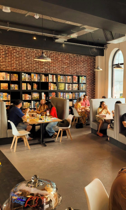 Inside The Dice Box Sheffield, there are tables and booths full of people playing games and drink and eating. The back wall is covered in shelving that is full of boxed games.
