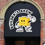 Builder's Brew Café logo on the outside of a building. The logo has the words 'Builder's Brew' round a cartoon character, who is shaped like a toy building brick, holding a mug of tea.