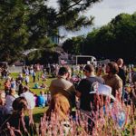 Shot looking towards the Devonshire Green main stage as people sit on the grass, framed by leaves from a tree above and some wild flowers 