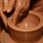 Close-up photograph of hands covered in wet clay shaping a ceramic pot on a spinning pottery wheel. The fingers are pressed inside the vessel to form its smooth, circular walls, while the clay rotates beneath them. Warm lighting highlights the glossy texture of the wet clay and the concentric ridges created during the pottery-making process.