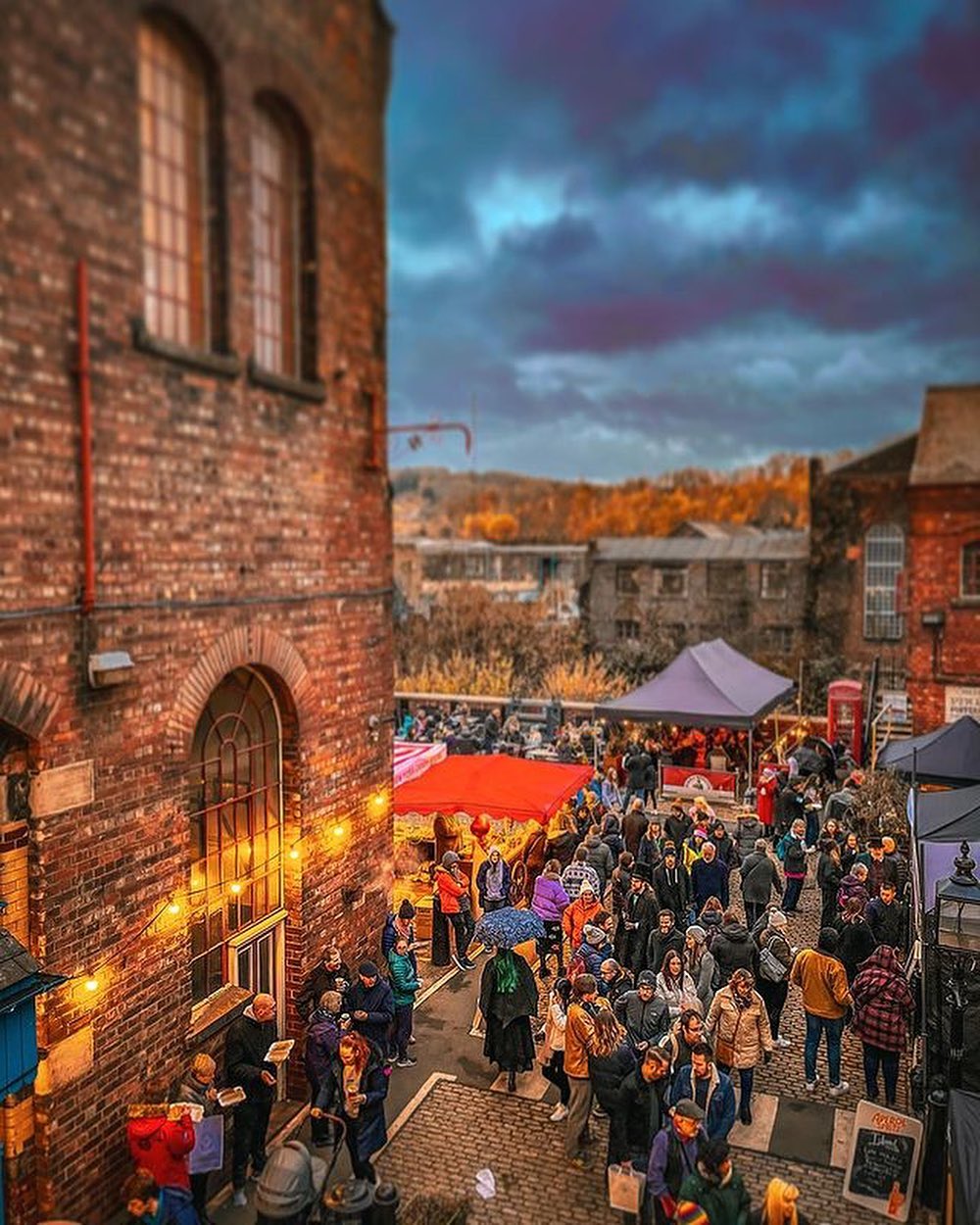 A bustling outdoor market set between historic brick buildings during twilight, with warm string lights illuminating the scene. Crowds of people walk along cobblestone paths, browsing stalls covered by colorful canopies, including bright red and purple tents. The background shows rolling hills and autumn trees under a dramatic, cloudy sky, creating a festive and vibrant atmosphere.