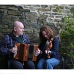 Two people seated outdoors beside a stone wall, playing traditional musical instruments. One person holds a wooden button accordion while the other plays a violin. A leafy potted shrub stands to one side, and the informal setting creates the feel of an intimate live music performance in a courtyard or garden space.