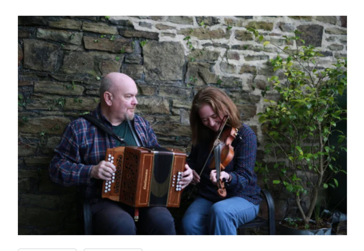 Two people seated outdoors beside a stone wall, playing traditional musical instruments. One person holds a wooden button accordion while the other plays a violin. A leafy potted shrub stands to one side, and the informal setting creates the feel of an intimate live music performance in a courtyard or garden space.