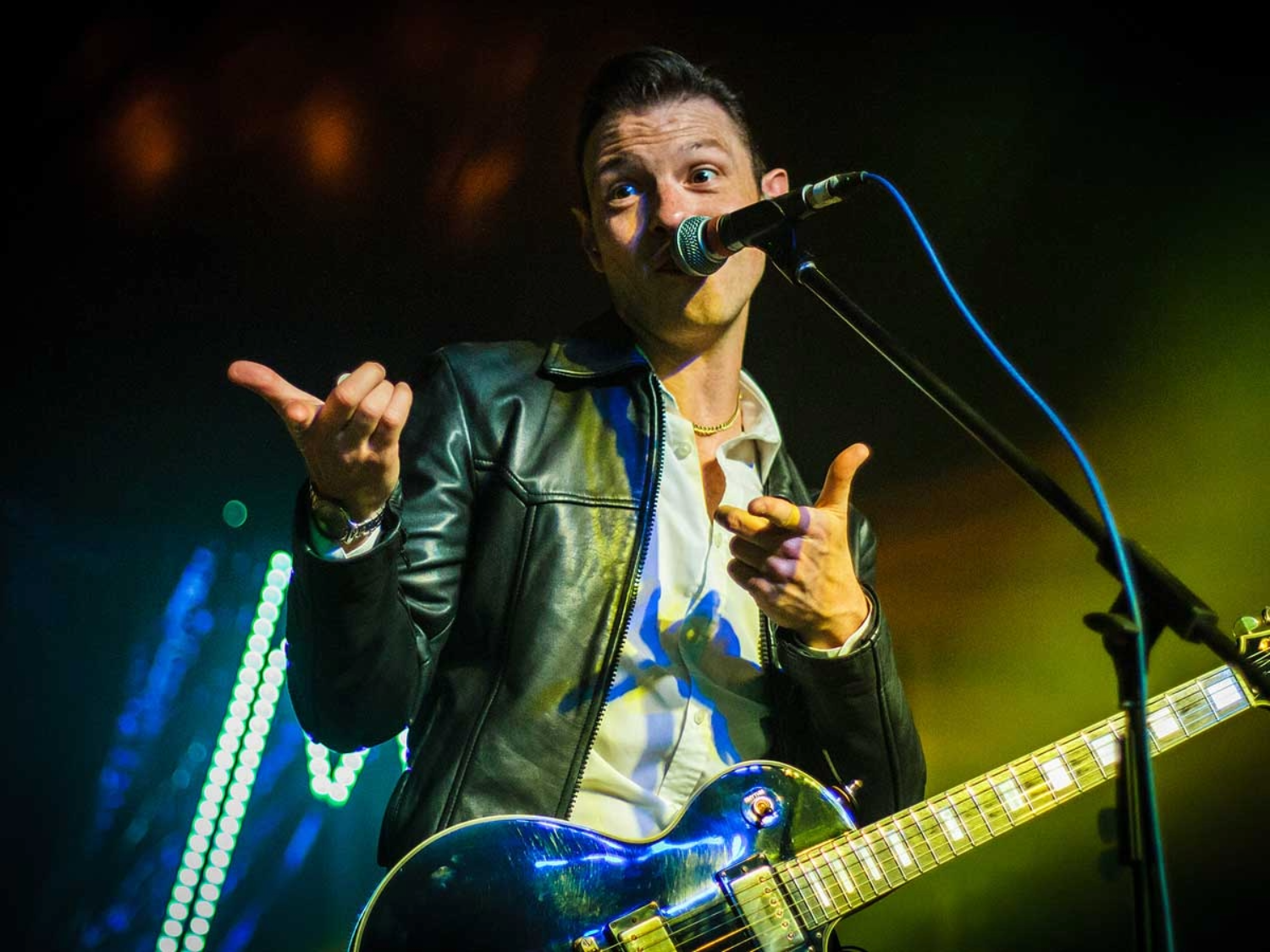 A musician on stage wearing a black leather jacket and white shirt, holding an electric guitar with a shiny blue finish. The performer stands in front of a microphone, gesturing with both hands as colourful stage lights create a vibrant backdrop with green and yellow tones.