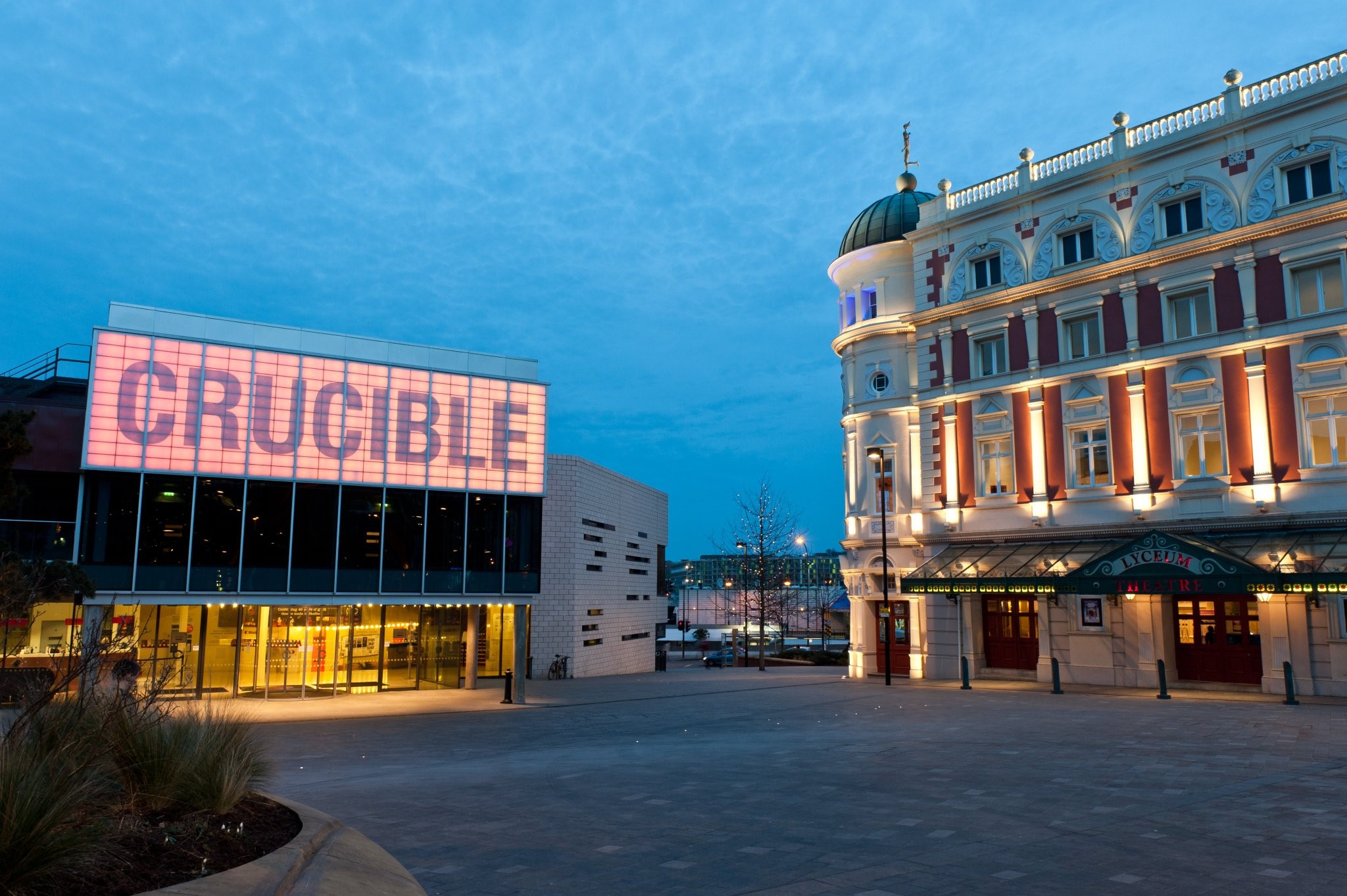 Evening view of Sheffield’s Crucible Theatre and the Lyceum Theatre. The Crucible building has a modern glass façade with large illuminated letters spelling “CRUCIBLE” in pink tones. To the right, the Lyceum Theatre features ornate architecture with columns, decorative details, and warm lighting highlighting its dome and entrance canopy. The sky is a deep blue, and the open plaza in front is quiet and spacious.