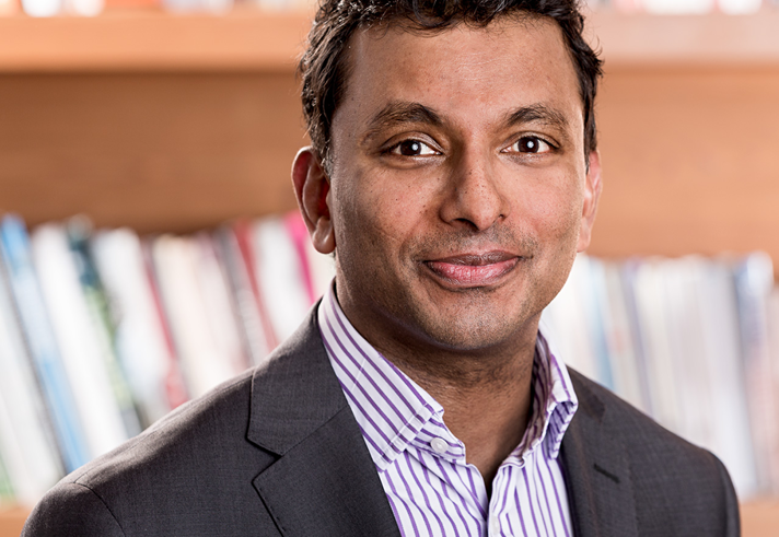 Person wearing a dark suit jacket over a light-colored, striped collared shirt, standing in front of a wooden bookshelf filled with books.
