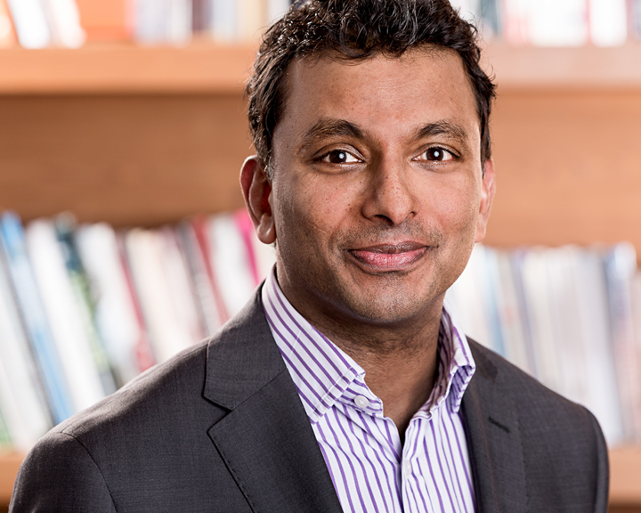 Person wearing a dark suit jacket over a light-colored, striped collared shirt, standing in front of a wooden bookshelf filled with books.