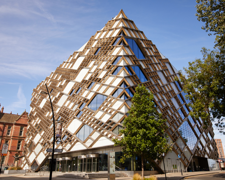 A modern, pyramid-shaped building with a striking geometric design featuring a mix of glass panels and wooden lattice structures. The facade reflects the blue sky, and the building is surrounded by trees and a few older brick buildings on the left. The entrance area has large glass doors, and the street in front includes signage and a lamppost.
