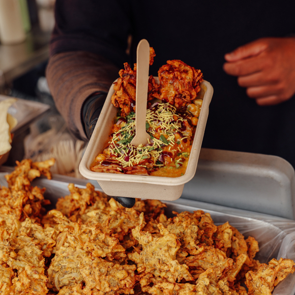 A tray of street food topped with sauce and herbs being held above a counter filled with fried items.