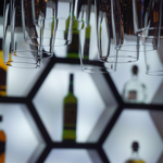 Close-up of an illuminated bar display featuring hexagonal shelves with bottles of liquor arranged inside. Several upside-down hanging glasses are in the foreground, creating a layered effect. The shelves have a bright white backlight, contrasting with the dark surroundings and highlighting the shapes of the bottles and glassware.