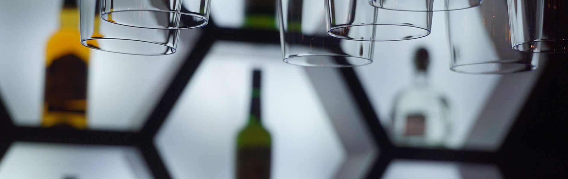 Close-up of an illuminated bar display featuring hexagonal shelves with bottles of liquor arranged inside. Several upside-down hanging glasses are in the foreground, creating a layered effect. The shelves have a bright white backlight, contrasting with the dark surroundings and highlighting the shapes of the bottles and glassware.
