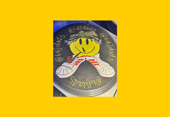Close-up of a customised DJ turntable slipmat featuring a large yellow smiley face wearing a patterned bucket hat, smoking a pipe, and resting on a pair of white trainers with red stripes. Handwritten yellow text curves around the design, reading “@6djdads” and “@6djdas”. The circular artwork sits on a black turntable platter with metallic edging, illuminated by blue-purple lighting.