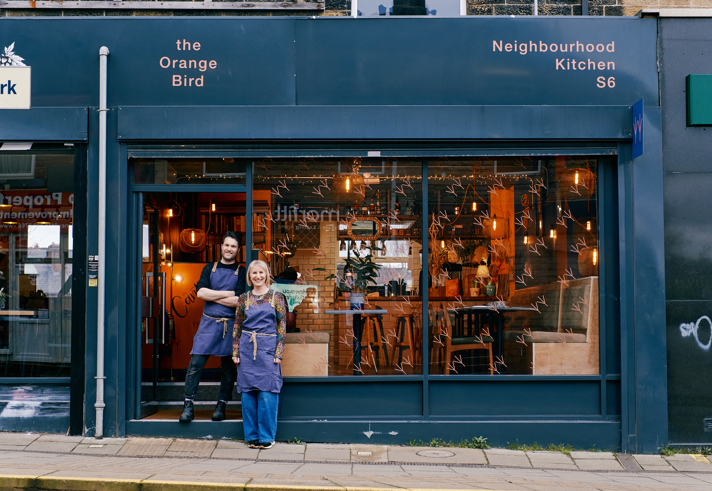 Front view of The Orange Bird restaurant at during the day with two friendly owners standing out front ready to welcome you 