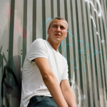 A person in a plain white T-shirt and jeans sits leaning against a corrugated metal wall covered with graffiti. The photograph is taken from a low angle, with the vertical lines of the grey metal panels stretching into the background. Bright daylight illuminates the scene, creating strong contrast between the white clothing and the industrial urban setting. The person looks off to one side while seated against the wall.