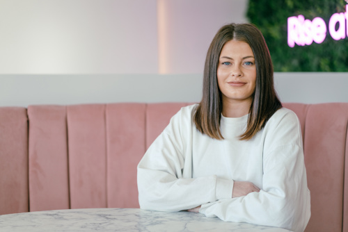 A person wearing a plain white long-sleeve top is seated at a round marble table with arms resting on the surface. The background features a pink cushioned booth and a wall with greenery and partially visible neon text. The setting appears modern and stylish, likely in a café or casual dining space.