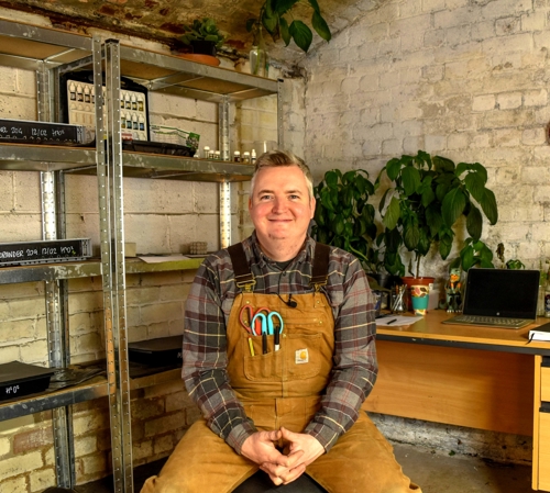 A man is sat, smiling, on a stool in a stone building.