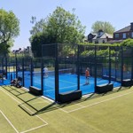 Outdoor padel courts with bright blue playing surfaces enclosed by glass and metal fencing, set within a larger green tennis court area. Several players are on the padel courts, holding rackets and playing under clear blue skies. Surrounding the courts are tall trees and residential houses, creating a leafy suburban backdrop. Sports bags and equipment are placed near the court entrance.