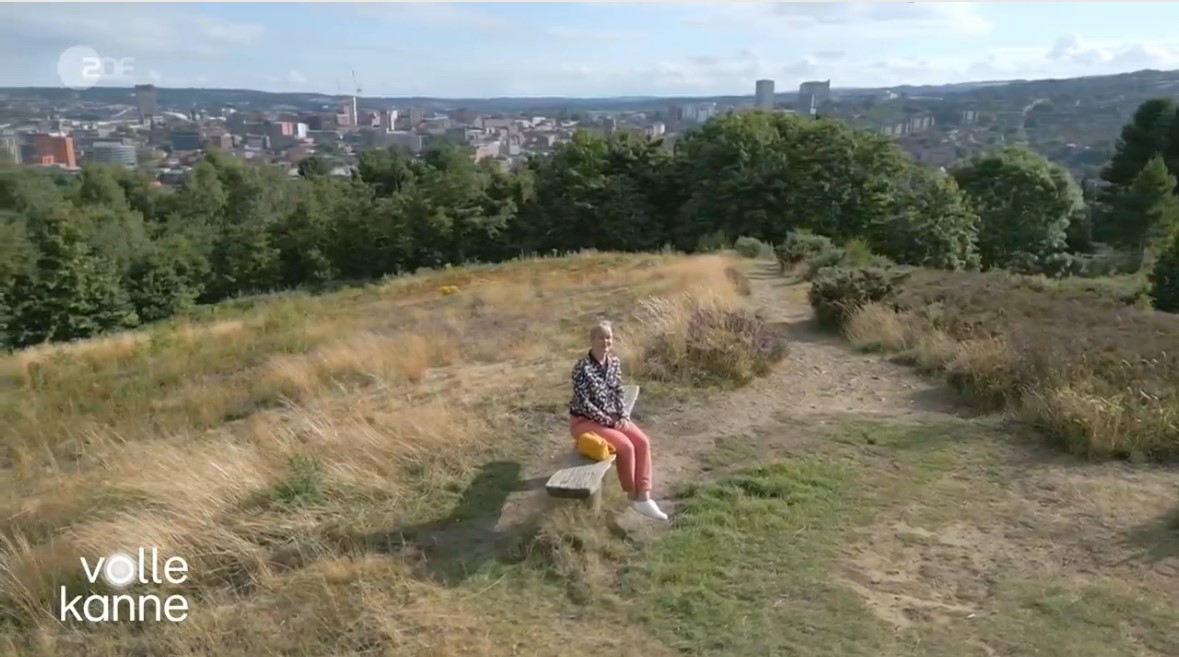 Person sitting on a stone bench along a grassy hillside path with patches of dry grass and shrubs, overlooking a cityscape in the distance under a partly cloudy sky. Text on the bottom left reads ‘volle kanne.’