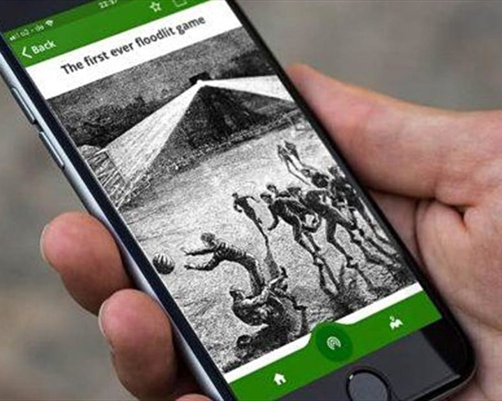 A hand holding a smartphone with the Home of Football: Walking Tour App on the screen.