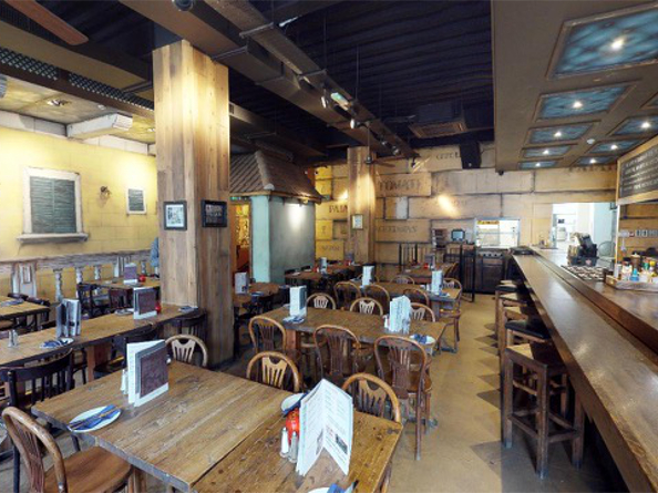The interior of Cubana Tapas Bar. Interior of a rustic-style restaurant featuring wooden tables and chairs arranged in rows, each set with menus and condiments. A long wooden bar with high stools runs along the right side, with a counter displaying various items. The walls have a textured, warm-toned finish with framed pictures and decorative elements. Overhead, dark exposed beams and recessed lighting create a cozy atmosphere.