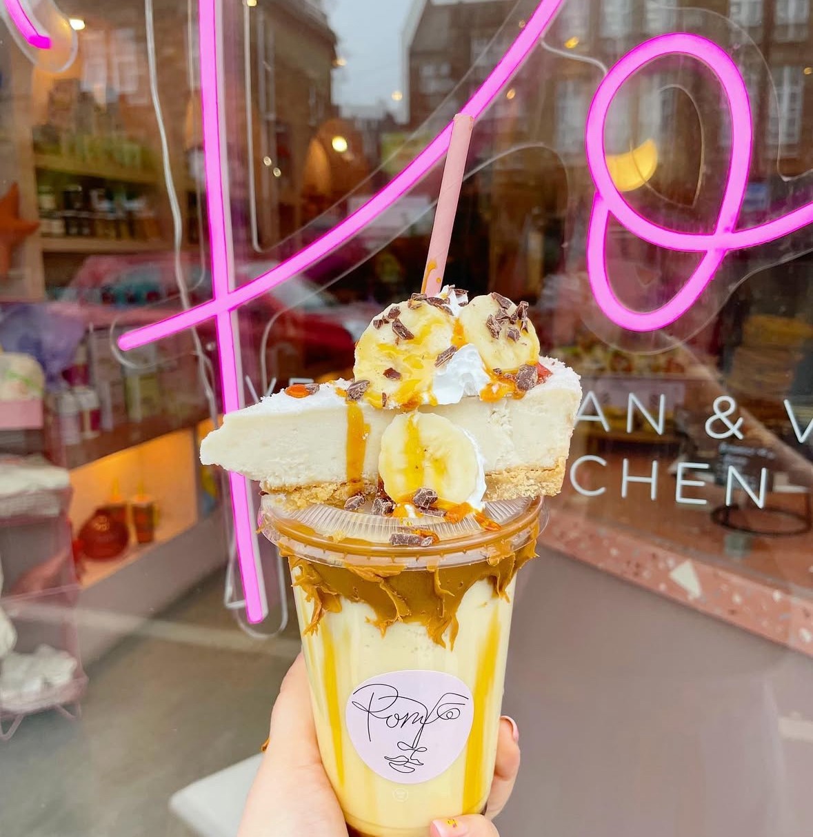 Hand holding a dessert drink topped with a slice of pie, banana slices, whipped cream, and caramel drizzle. The drink is in a clear cup with a pink straw and a label reading ‘Pom.’ In the background, a shop window with pink neon signage and shelves of colorful items is visible.